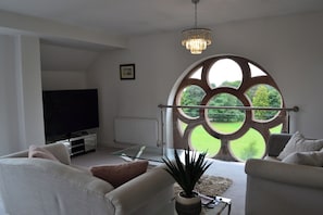 Una sala de estar moderna con un televisor grande, asientos cómodos, una mesa de centro de vidrio y una vista a través de una ventana peculiar.