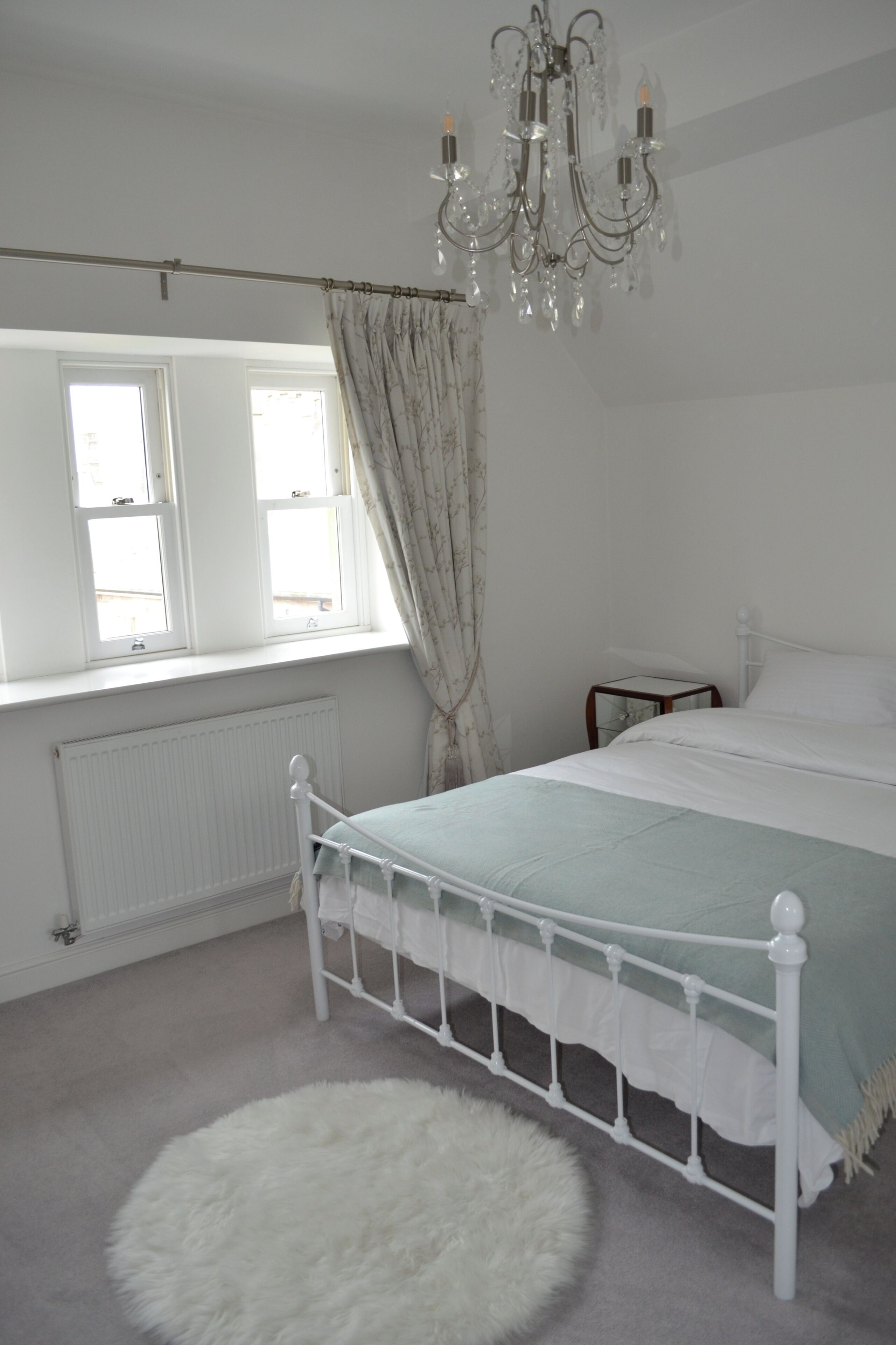 A bedroom with a white bed, a radiator, a window with curtains, and a chandelier.