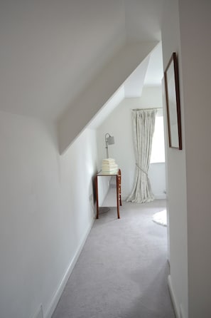 A narrow hallway with a sloped ceiling, a small table with a lamp, and a window with curtains. - The Residence, 3-bed Apt, Sleeps 6 With Parking (Lancaster)
