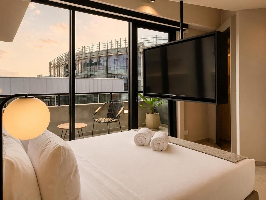 A modern hotel room with a large bed, a flat-screen TV, a balcony with a view of a building under construction, and a potted plant.