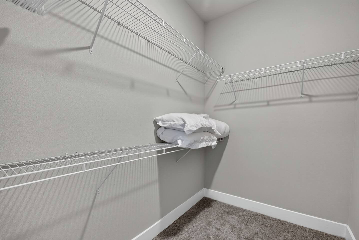 A corner room with a white carpet, a white wall, and a metal wire shelf with folded white linens.