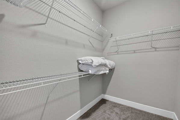 A corner room with a white carpet, a white wall, and a metal wire shelf with folded white linens. - Plush 2BR Apt/Pool/Gym/King Bed/Beachwalk access (St. Johns County)