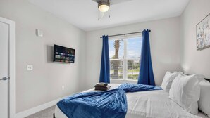 Ein Schlafzimmer mit einem Bett, einem Deckenventilator, einem Fenster mit Vorhängen und einem an der Wand befestigten Fernseher.