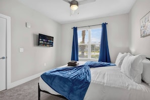 Ein Schlafzimmer mit einem Bett, einem Deckenventilator, einem Fenster mit Vorhängen und einem an der Wand befestigten Fernseher.