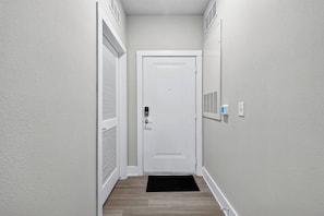 Ein schmaler Flur mit einer weißen Tür, einer schwarzen Fußmatte und einem an der Wand befestigten Thermostat.