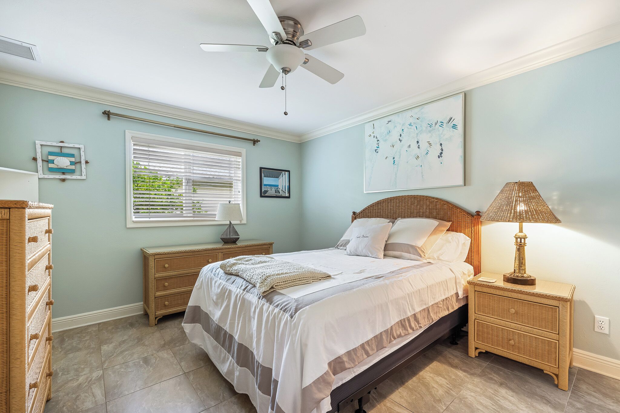 Une chambre à coucher comprenant un lit, une table de chevet, une lampe, un ventilateur de plafond et une fenêtre avec des stores.