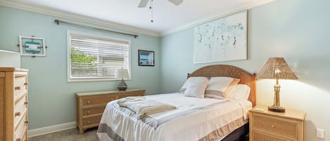 Een slaapkamer met een bed, nachtkastje, lamp, plafondventilator en raam met jaloezieën.