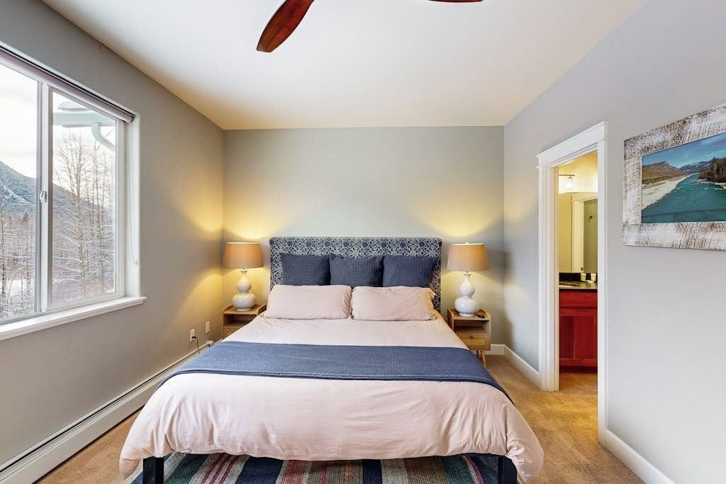 A bedroom with a bed, bedside lamps, a window, and a painting on the wall.