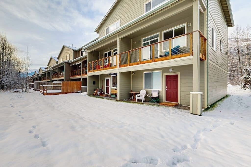 A multi-story residential building with a balcony on the second floor, a red door, and a small outdoor seating area.