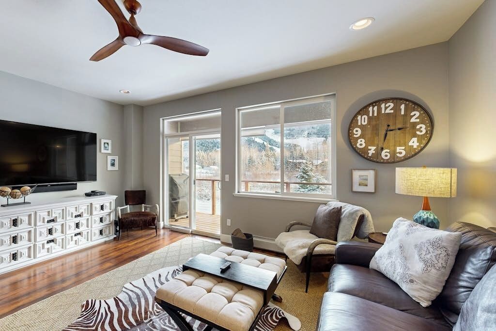 A living room with a large flat-screen TV, a ceiling fan, a large wall clock, and a sofa with a patterned throw pillow.