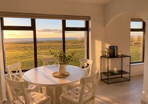 A dining area with a round table and chairs, a vase with flowers, and a view of a grassy field through large windows.