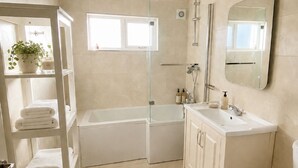Un baño moderno con bañera, inodoro, lavamanos y ducha. Hay un estante con toallas y una planta en maceta.