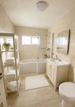 A modern bathroom with a bathtub, a toilet, a sink, and a shower. There is a shelf with towels and a potted plant.
