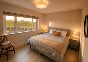 Ein Schlafzimmer mit einem großen Bett, zwei Nachttischen, einem Sessel und einem Fenster mit Blick auf ein Wiesenfeld.