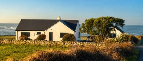 A coastal house with a stone wall, a grassy foreground, and a view of the sea.
