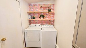A laundry room with two white washing machines, a pink patterned wall, and a shelf with books and plants.