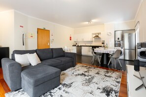 Un appartement moderne doté d’un canapé d’angle gris, d’un coin repas, d’une cuisine équipée d’appareils électroménagers en acier inoxydable et d’un petit espace buanderie.