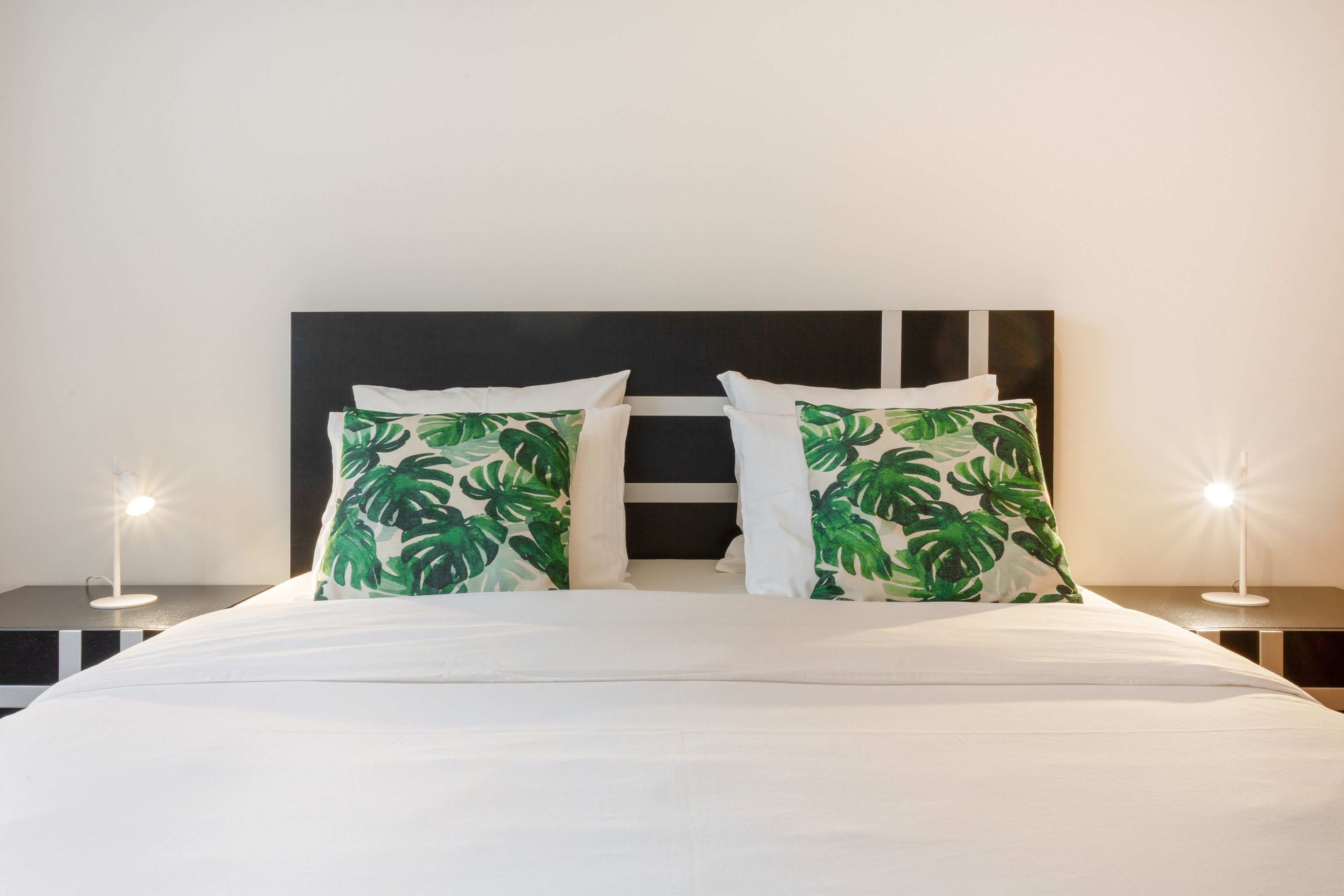 Una cama con ropa de cama blanca y dos almohadas con estampado de hojas verdes, flanqueada por dos mesitas de noche con lámparas.