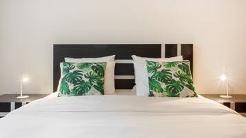 Una cama con ropa de cama blanca y dos almohadas con estampado de hojas verdes, flanqueada por dos mesitas de noche con lámparas.