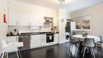 A modern kitchen with white cabinetry, a dishwasher, an oven, and a washing machine. There is a dining table with chairs and a window with a view of the outdoors.