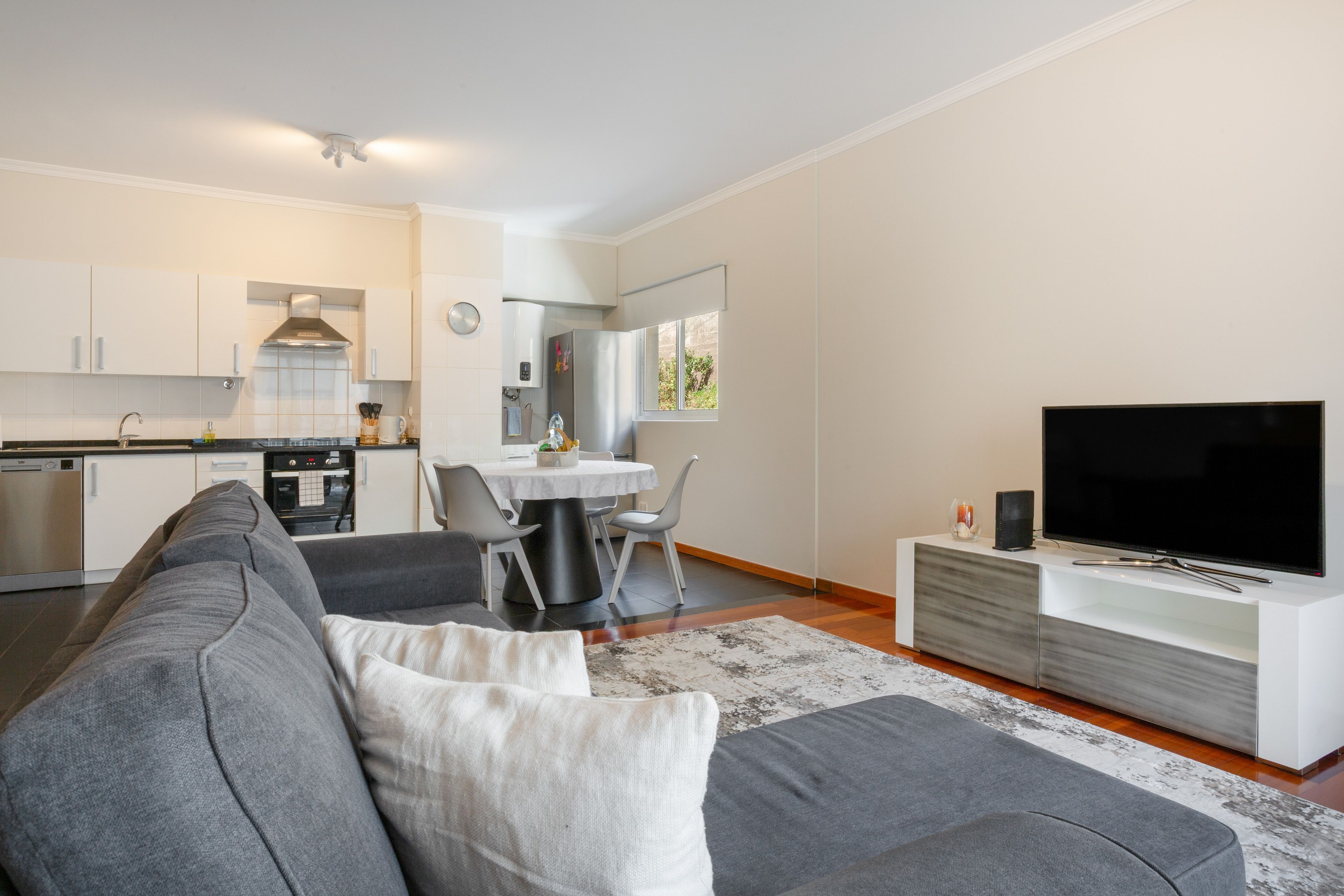 Een moderne woonruimte met een grijze bank, een flatscreen televisie en een eethoek met een tafel en stoelen.