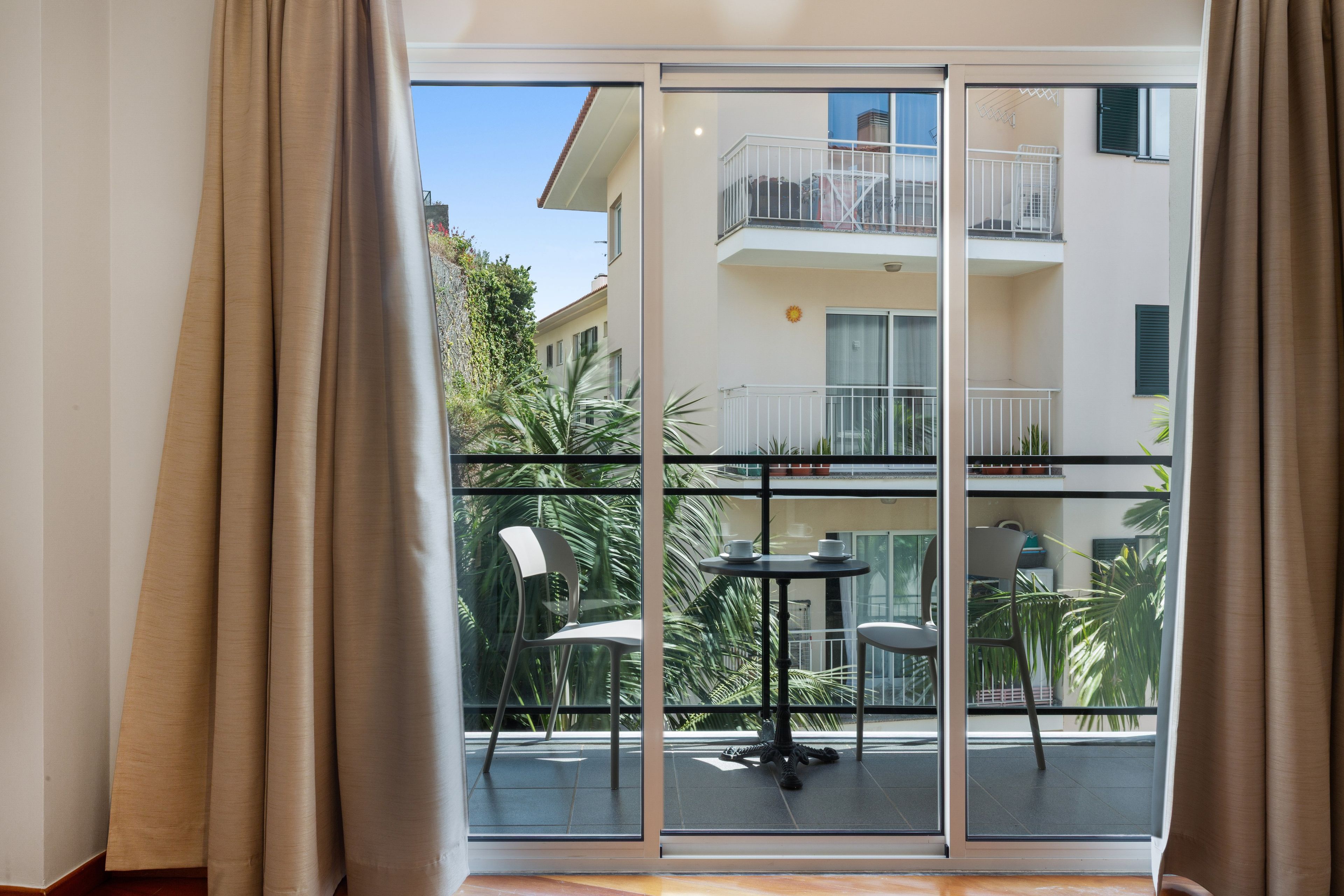 Una habitación con puerta corrediza de vidrio que da a un balcón con mesa y sillas, y vista a un edificio con balcones.