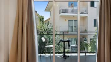 Una habitación con puerta corrediza de vidrio que da a un balcón con mesa y sillas, y vista a un edificio con balcones.