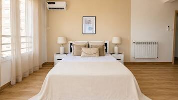 Une chambre à coucher avec un lit, deux tables de chevet avec des lampes, un climatiseur fixé au mur, un tableau encadré et un radiateur.