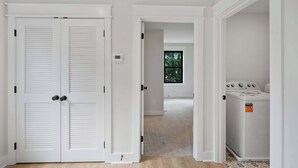 Een kamer met een witte deur, een raam met uitzicht op bomen, en een wasmachine.