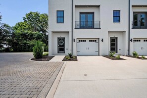 A modern two-story building with a white facade, a front entrance with a door, and two garage doors. - Rooftop Retreat: Steps to Downtown Square (Murfreesboro)