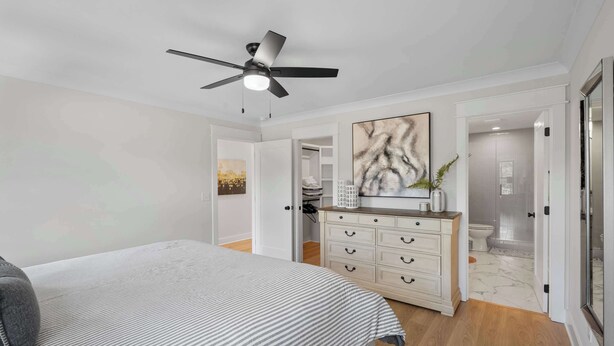 Een slaapkamer met een bed, een dressoir, een plafondventilator en een zichtbare badkamer door een geopende deur.