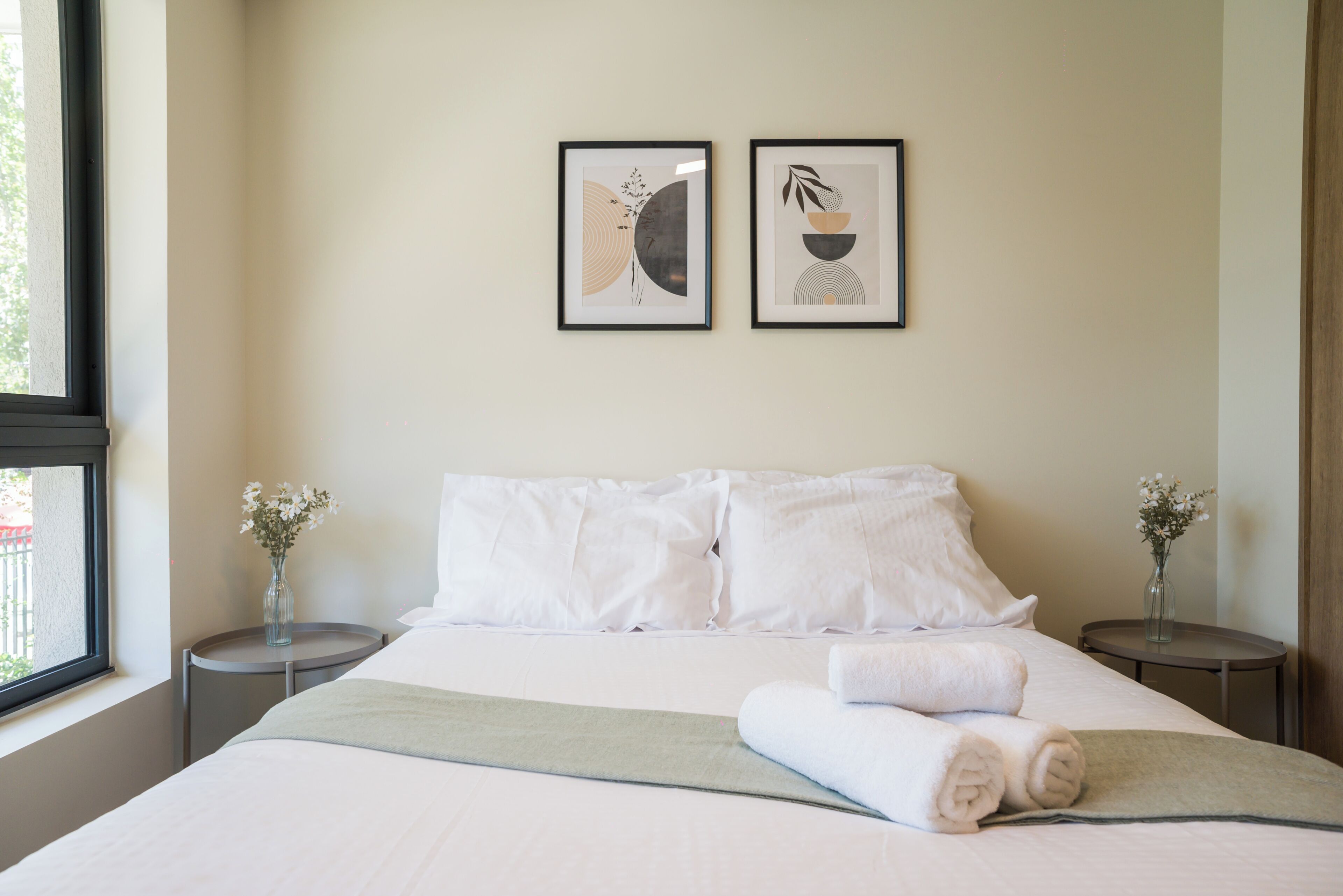 Quarto de hotel com uma cama, duas mesas de cabeceira, uma janela e duas obras de arte emolduradas na parede.