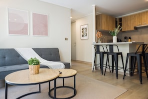 Een moderne woonkamer met een grijze bank, twee ronde tafels en een keukenhoek met houten kasten en barstoelen.