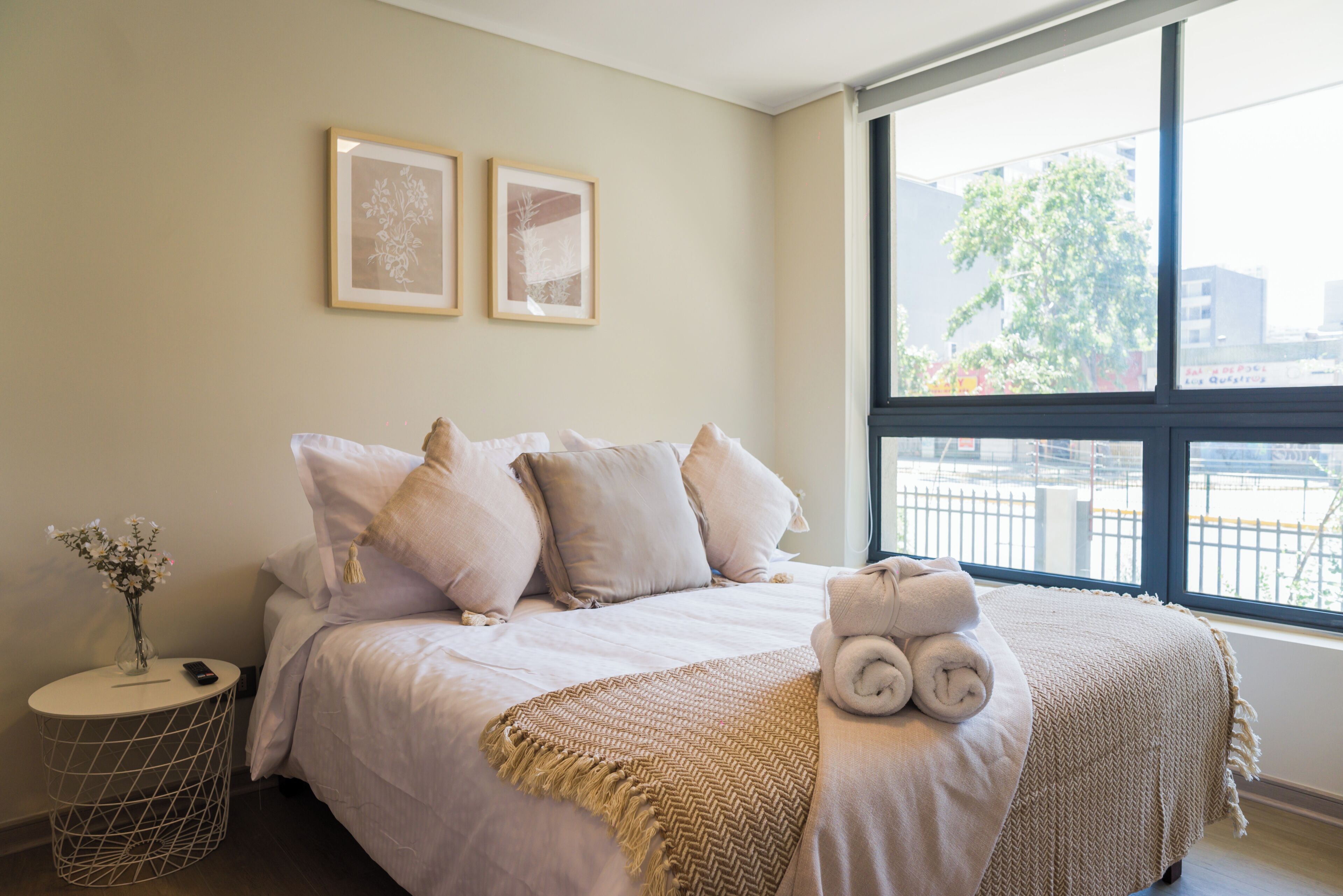 A bedroom with a bed, pillows, a bedside table with a vase, and a window with a view of buildings.