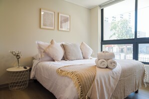 A neatly made bed with a beige blanket and pillows, a small round table with a vase of flowers, and a window with a view of buildings and trees. - Porvenir Apartments by Andes. Parking. (Santiago)