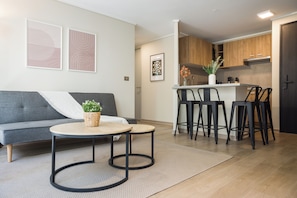 Un espacio de sala moderno con un sofá gris, una mesa de centro redonda y una mesita auxiliar con una planta. Hay tres taburetes negros y una zona de cocina con armarios de madera y encimera blanca.