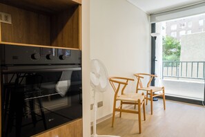 A kitchen with a stove, oven, and a white wall fan. There are two wooden chairs and a view of a balcony with a railing. - Porvenir Apartments by Andes. Parking. (Santiago)
