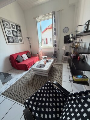 Ein Wohnzimmer mit einem roten Sofa, einem gemusterten Teppich, einem Couchtisch, einem Fernseher auf einem Regal und Blick ins Freie.