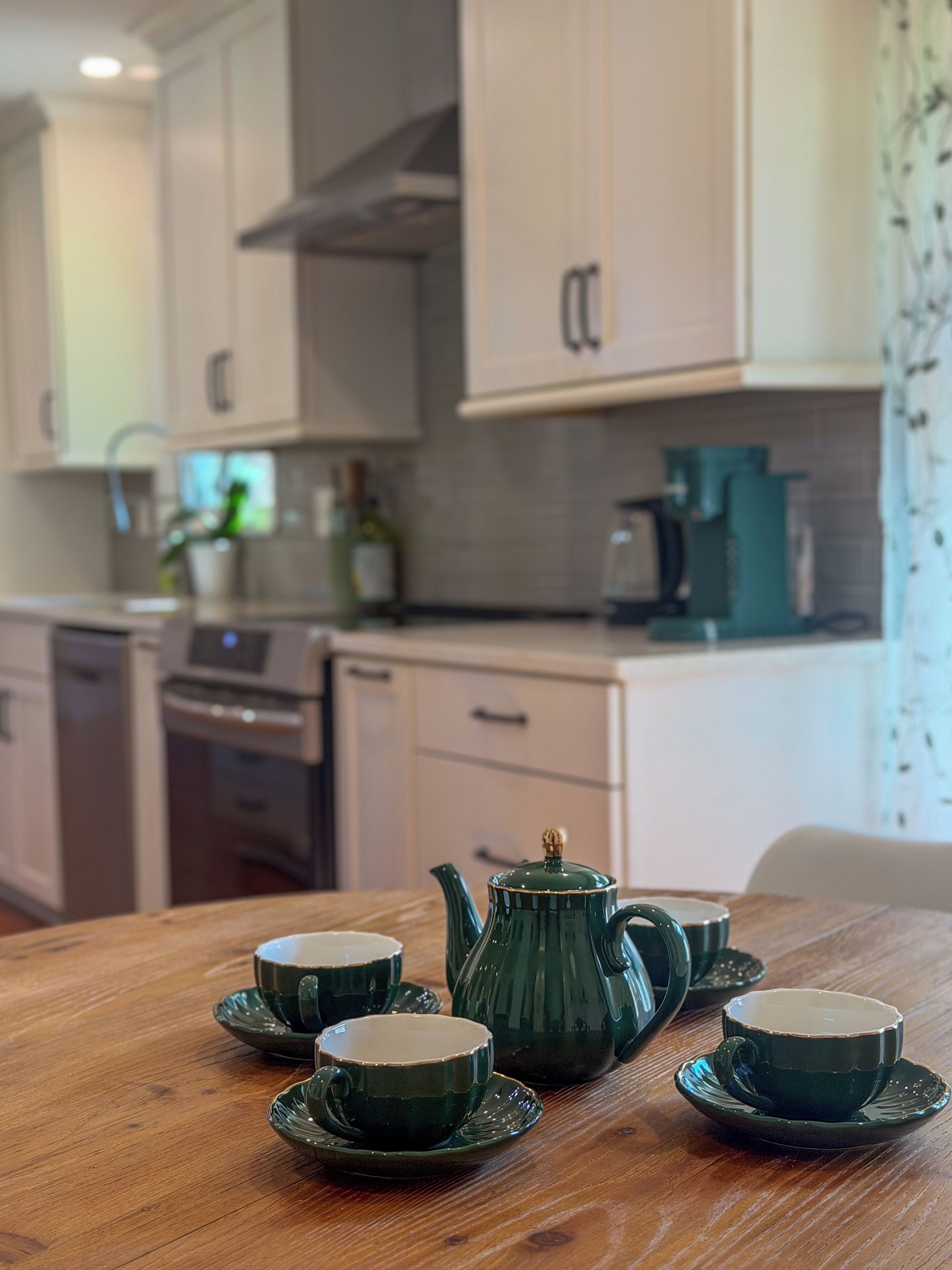 A wooden table with four green teapots and cups, a coffee maker, and a kettle in a kitchen with light-colored cabinets and a window with curtains.