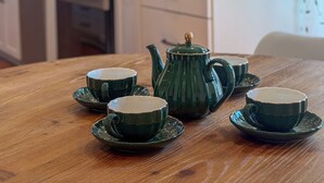 Ein Holztisch mit vier grünen Teekannen und Tassen, einer Kaffeemaschine und einem Wasserkocher in einer Küche mit hellen Schränken und einem Fenster mit Vorhängen.