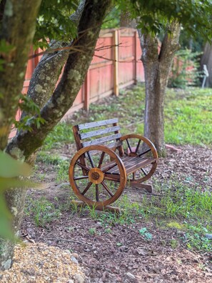 A wooden bench with large wheels, positioned under a tree in a garden area with grass and mulch ground cover. - Starling House/Huge Fenced Yard/Pet-Friendly (Mableton)