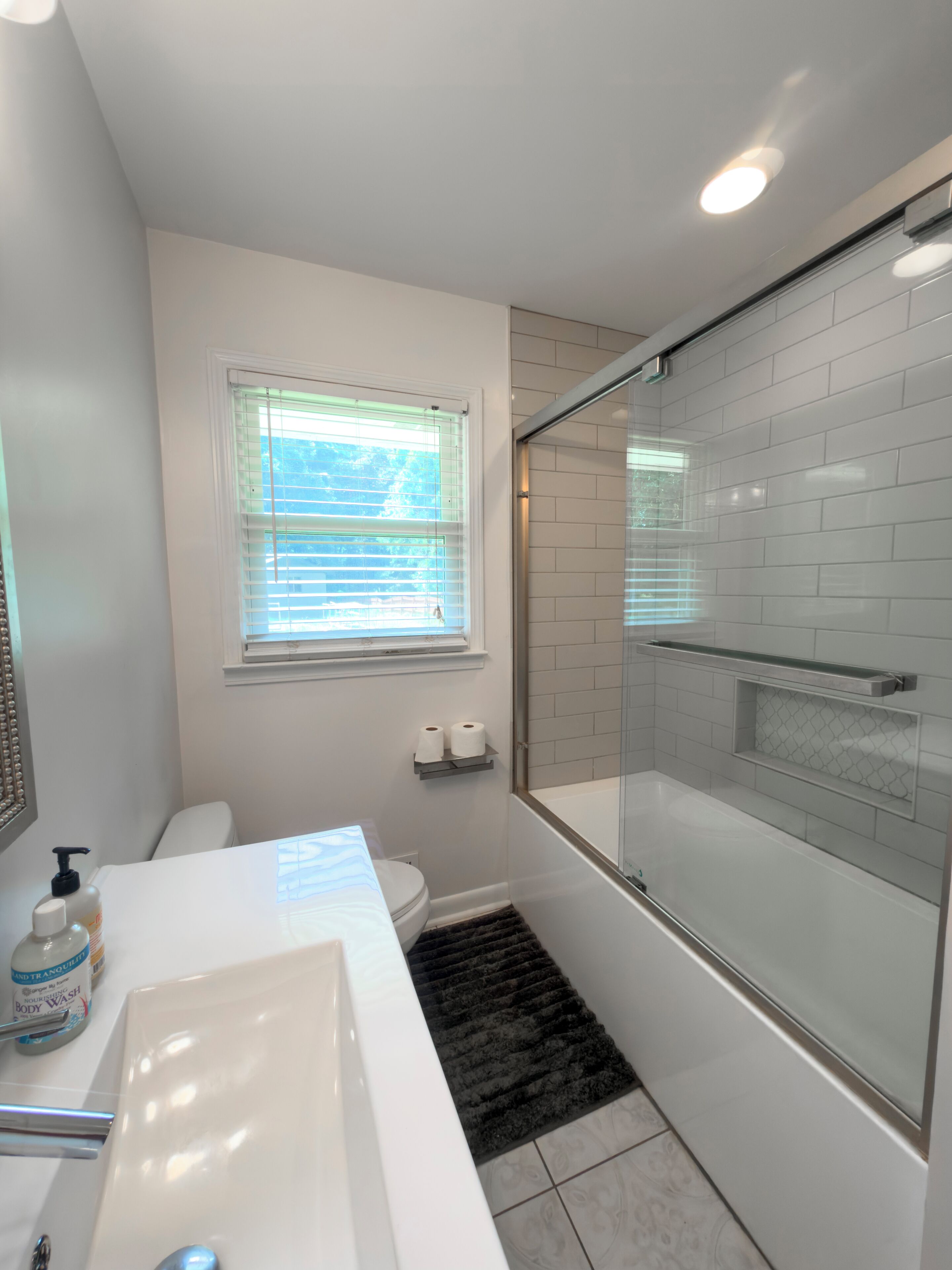 A bathroom with a white bathtub, a glass-enclosed shower, a white toilet, and a window with blinds.