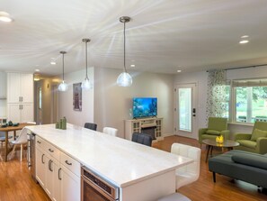 A modern kitchen with a central island, white cabinetry, and a built-in oven. There are pendant lights, a flat-screen TV, and a seating area with a sofa and chairs. - Starling House/Huge Fenced Yard/Pet-Friendly (Mableton)