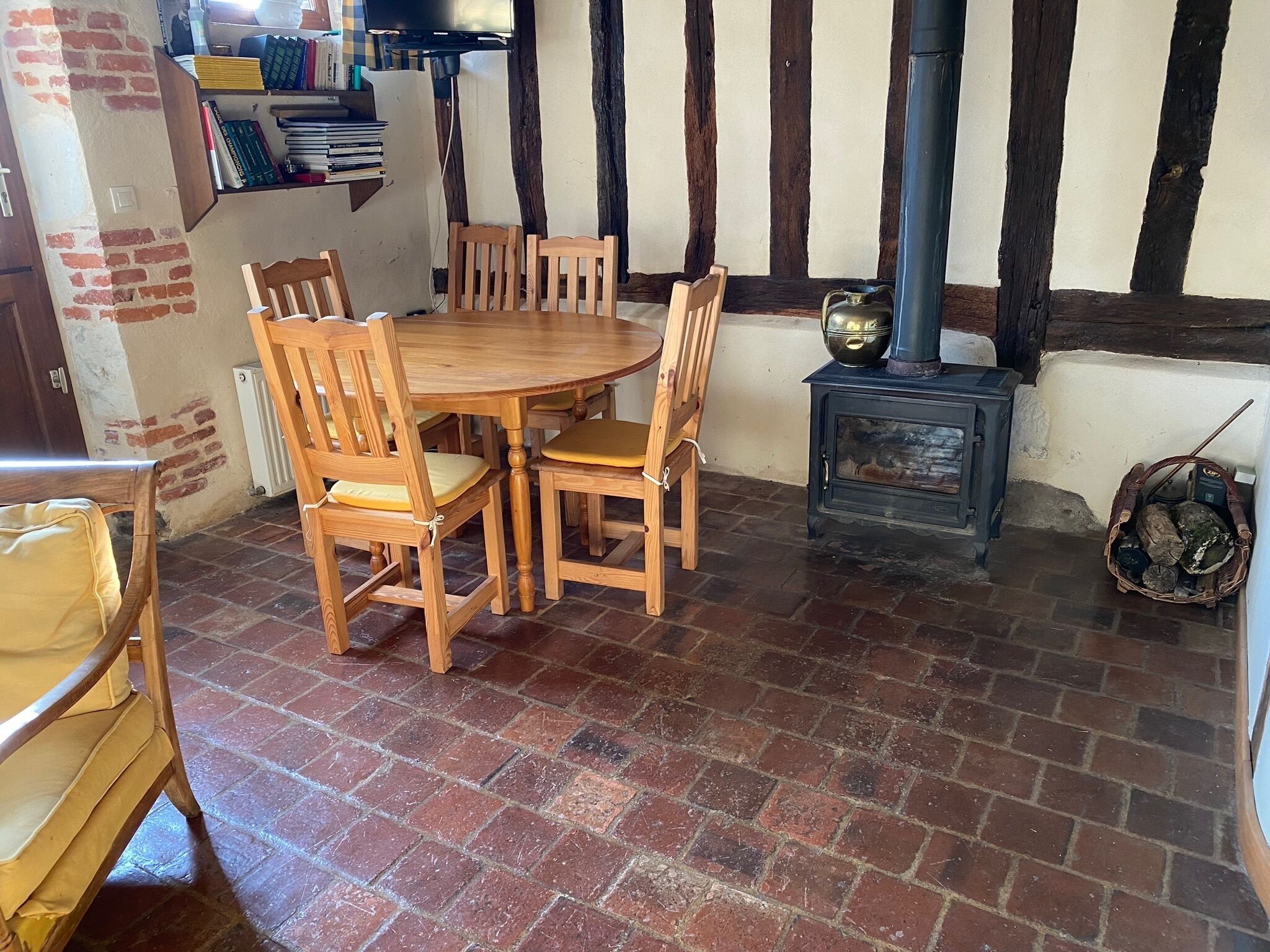 Une pièce à l’ambiance traditionnelle, avec une table et des chaises en bois, un sol en briques, une cheminée avec des bûches et une bibliothèque remplie de livres.