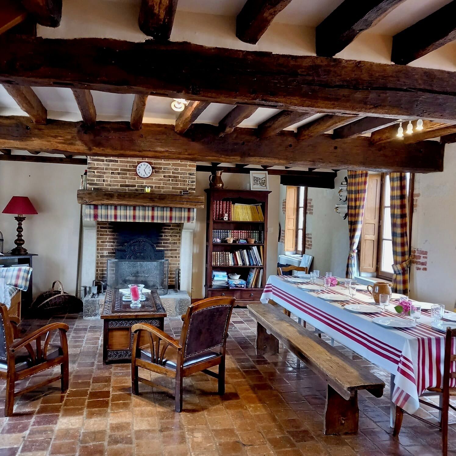 A rustic interior with exposed wooden beams, a brick fireplace, a dining table set for two, and a bookshelf.