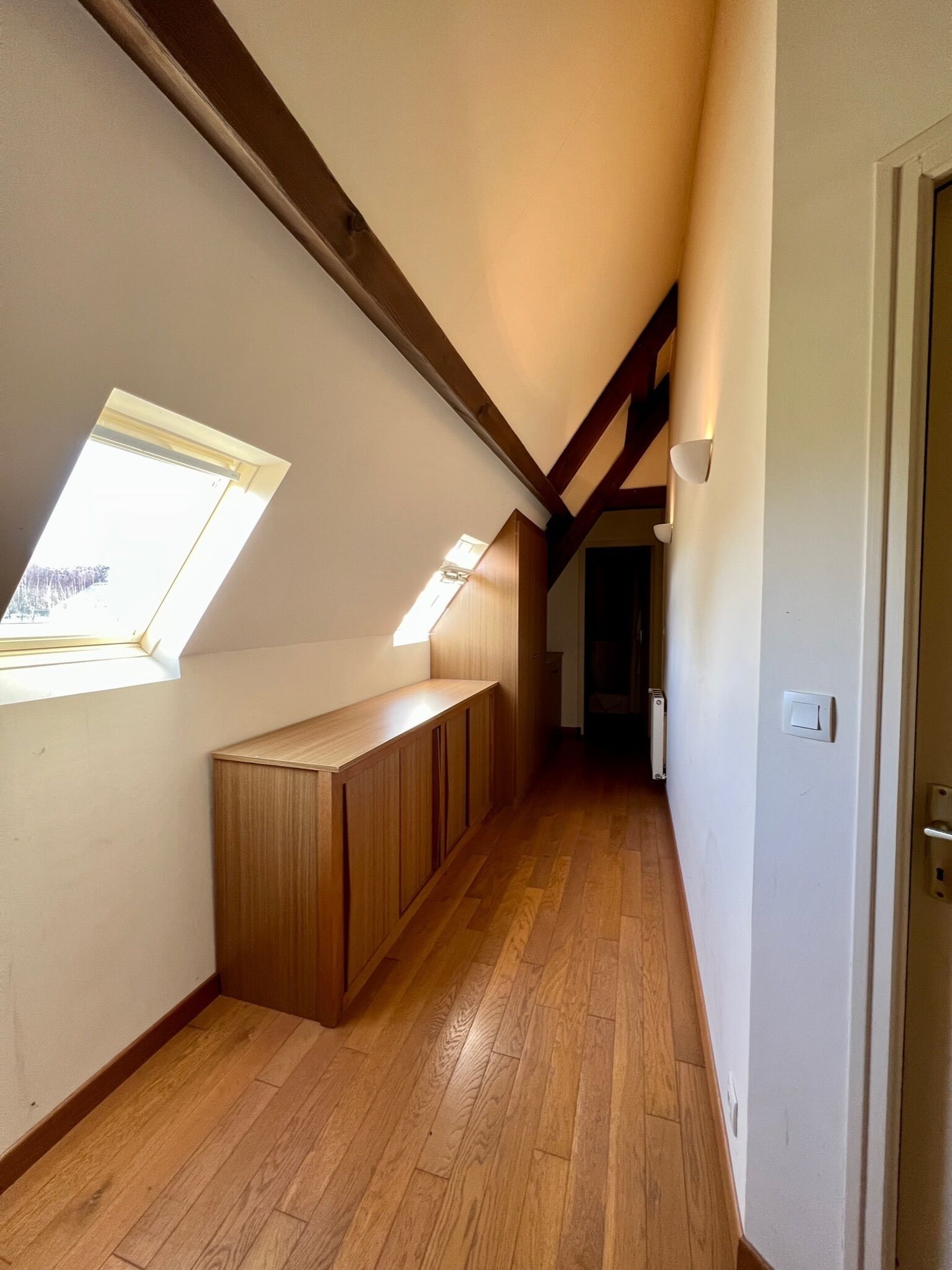 Un couloir étroit au plancher en bois, un plafond en pente doté de lucarnes et une porte sur le côté droit.