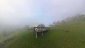 Een koepelvormige constructie met een houten platform, omgeven door een mistig landschap en groen.