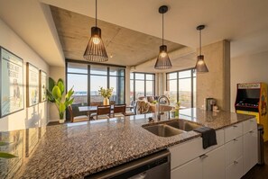 Une cuisine moderne dotée de plans de travail en granit, d’un éclairage suspendu et d’une grande fenêtre donnant sur un paysage urbain.