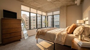 Une chambre à coucher comprenant un lit, une commode, un téléviseur et une fenêtre donnant sur des immeubles.