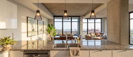 A modern kitchen with a granite countertop, stainless steel appliances, pendant lighting, and a view of the city through large windows.